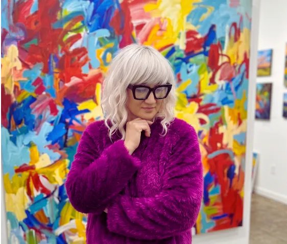 Kay in a magenta sweater and glasses, standing in front of a colorful abstract painting